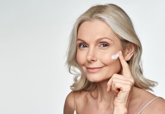 A woman with fair skin is shown smiling in a close-up photograph. She appears to be in her 60s, but her skin looks much younger and smoother than her age suggests. 