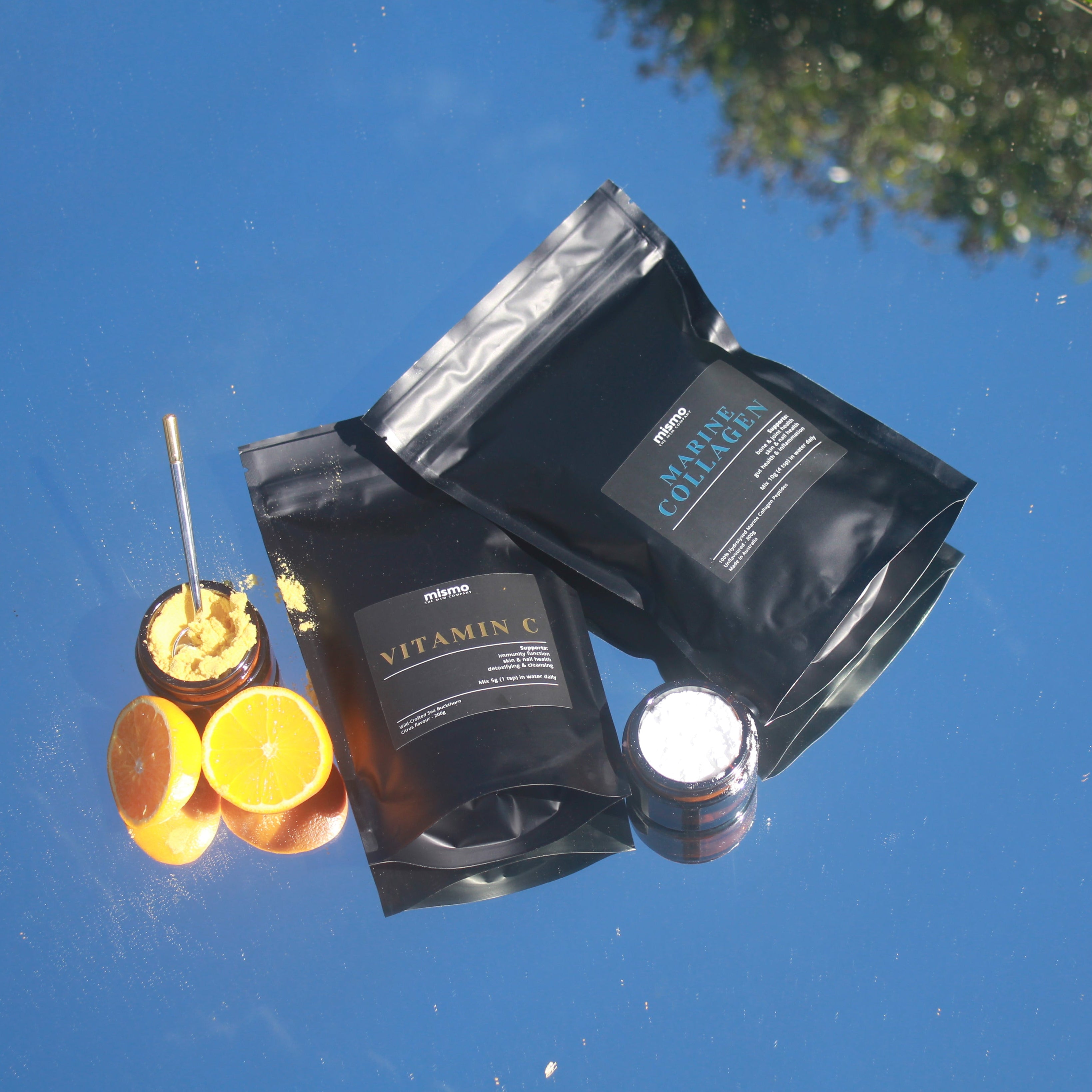 Three black pouches of skincare products labeled Vitamin C, Glycolic Acid, and Hyaluronic Acid, arranged on a highly reflective blue surface with orange slices and a small jar nearby.