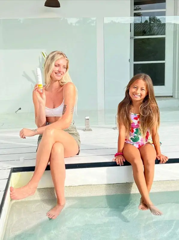 A young woman with blonde hair is sitting next to a swimming pool and holding an airless pump of tinted mineral sunscreen with an SPF of 50. The woman is wearing a swimsuit and is looking away from the camera with a smile. The woman's sitting next to a young girl indicating the sunscreen is safe for children. 