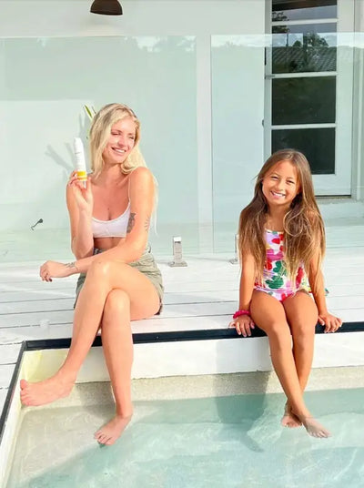 A young woman with blonde hair is sitting next to a swimming pool and holding an airless pump of tinted mineral sunscreen with an SPF of 50. The woman is wearing a swimsuit and is looking away from the camera with a smile. The woman's sitting next to a young girl indicating the sunscreen is safe for children. 